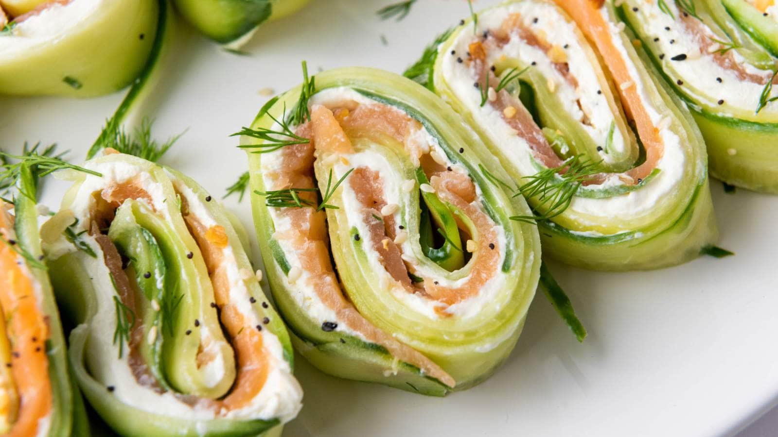 Close-up of cucumber roll-ups filled with cream cheese, smoked salmon, and herbs, garnished with fresh dill and black sesame seeds on a white plate.