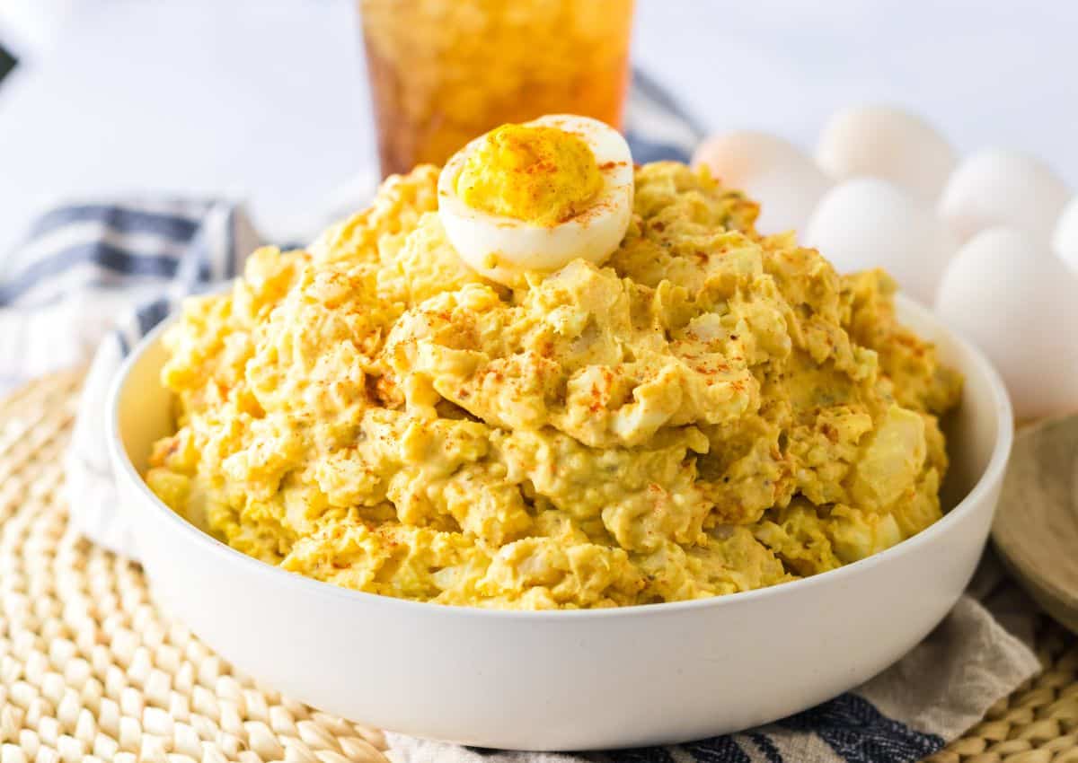 A bowl of potato salad topped with half a boiled egg and sprinkled with paprika, with a drink and whole eggs in the background.