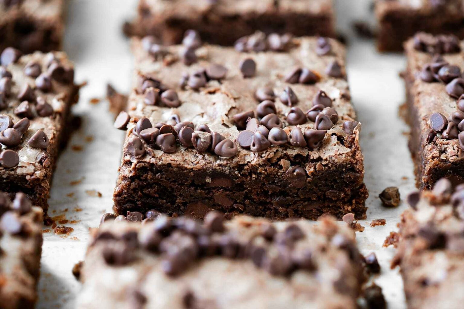 Close-up of chocolate chip brownies cut into rectangular pieces, topped with chocolate chips and arranged on a light-colored surface.