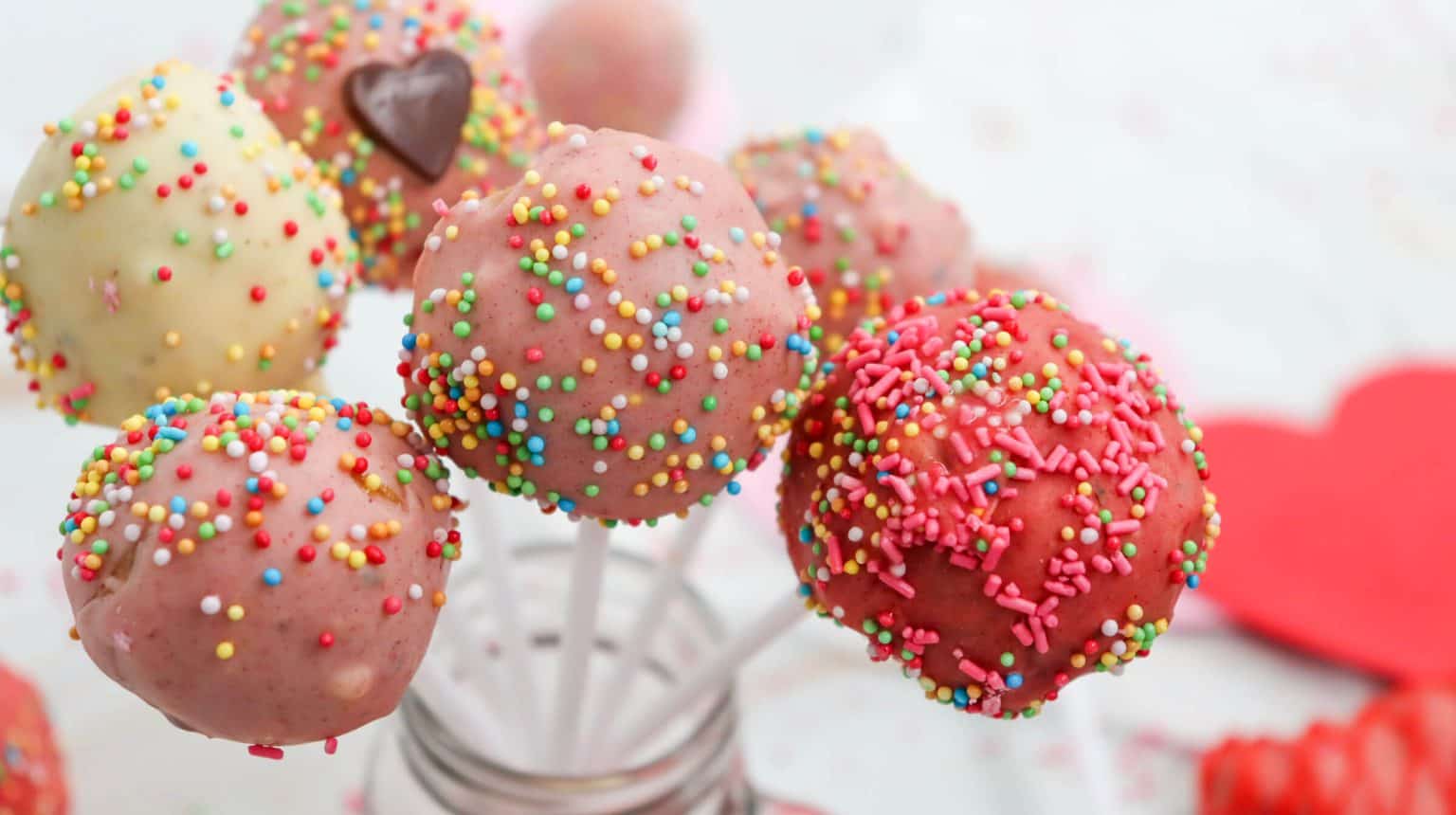 Colorful cake pops coated with pink, red, and white icing and decorated with sprinkles are arranged on sticks in a glass jar.
