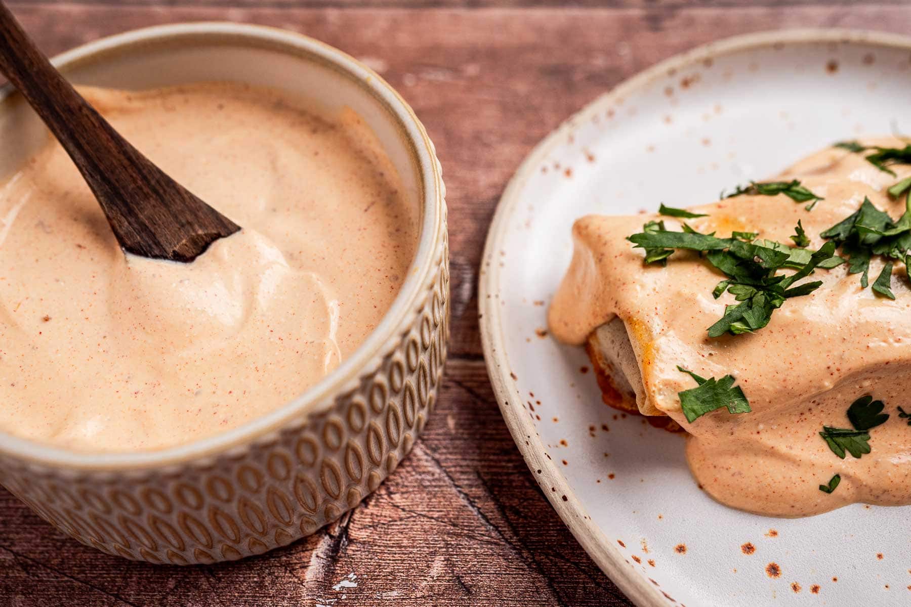 A bowl of creamy sauce with a wooden spoon next to a plate of food topped with the same sauce and garnished with chopped herbs.