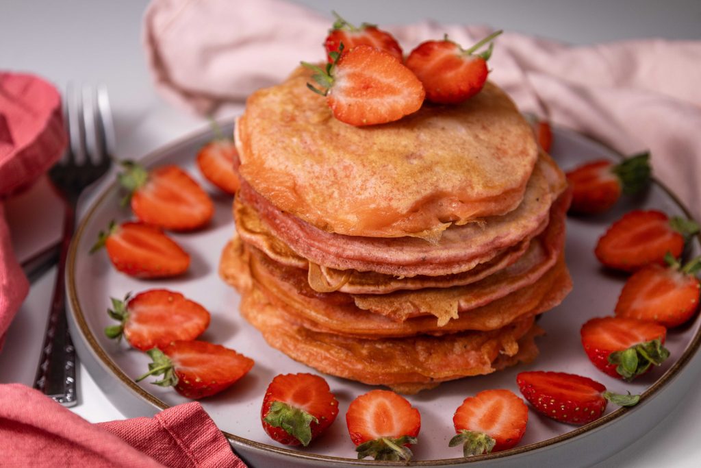 A stack of pancakes topped and surrounded by halved strawberries on a plate with a pink napkin and fork beside it.