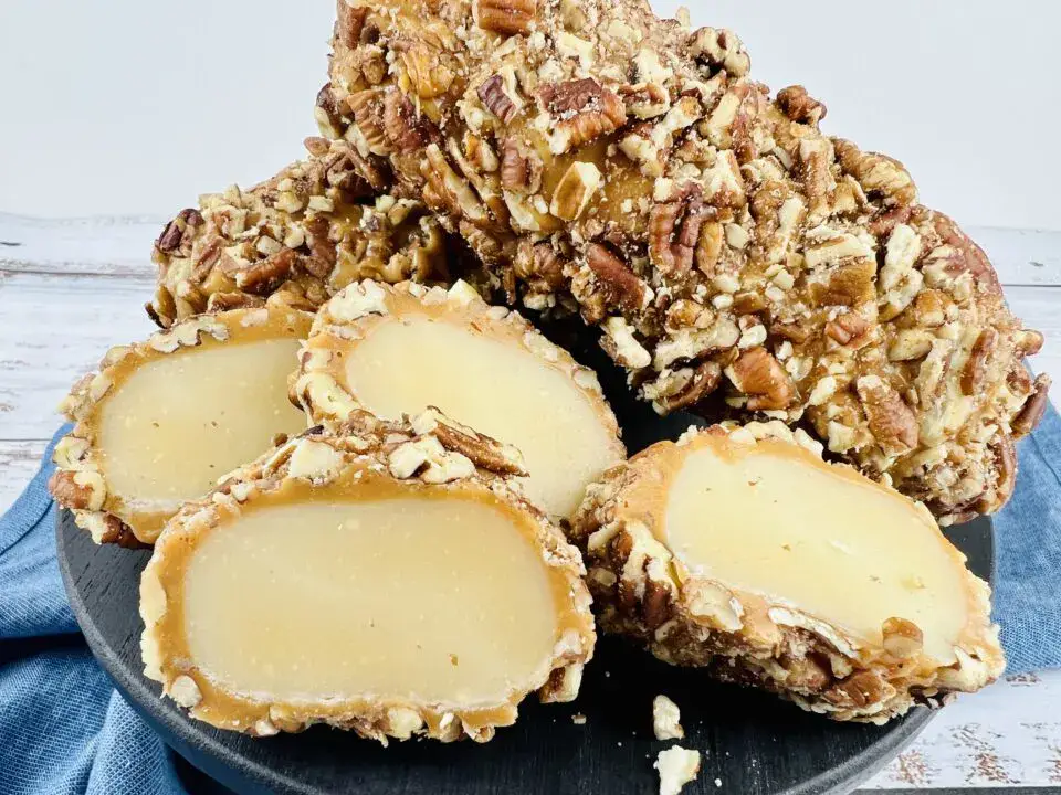 A black plate holds whole and sliced pecan-coated caramel candies with a creamy filling, displayed on a blue cloth with a light wooden background.