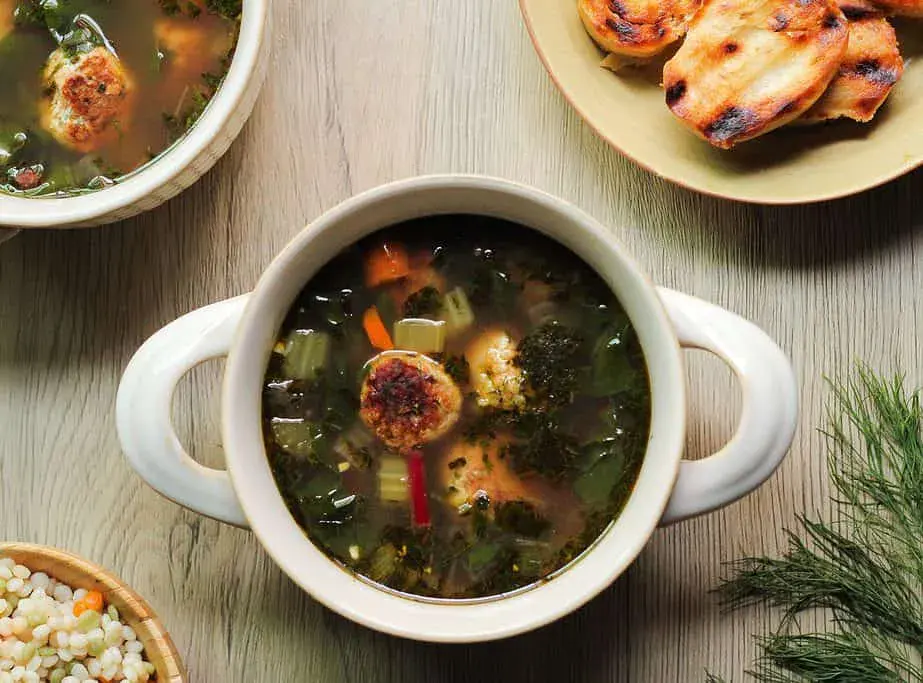 A bowl of vegetable soup with meatballs, surrounded by a plate of toasted bread, another bowl of soup, and a bowl of cooked grains on a wooden table.