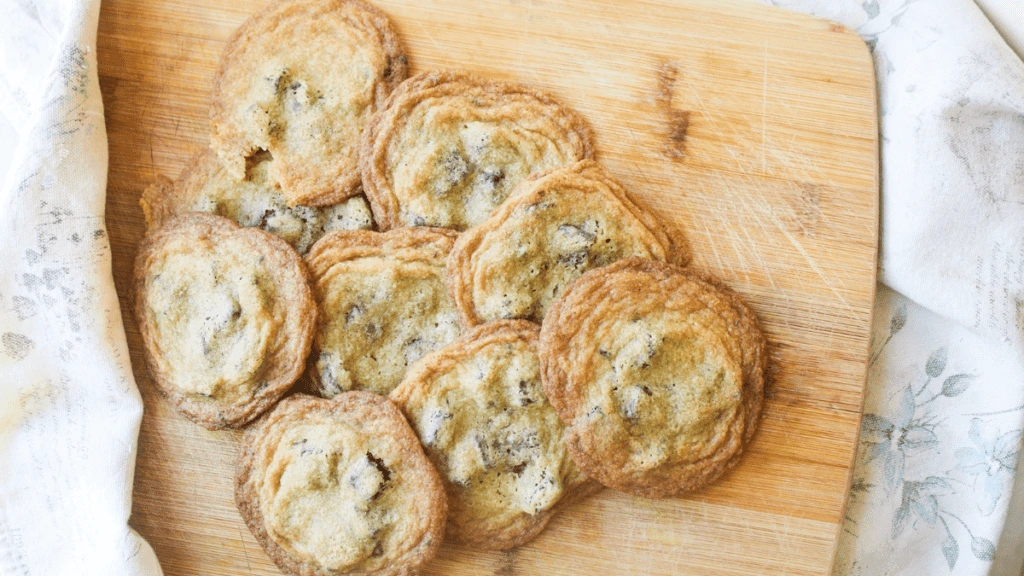 A group of chocolate chip cookies arranged on a wooden cutting board, partially bordered by a white cloth.
