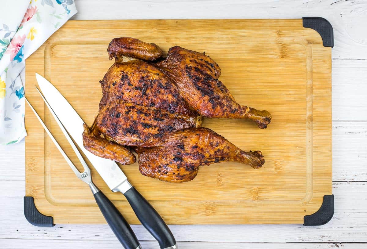 A grilled spatchcocked chicken rests on a wooden cutting board next to a chefs knife and carving fork.