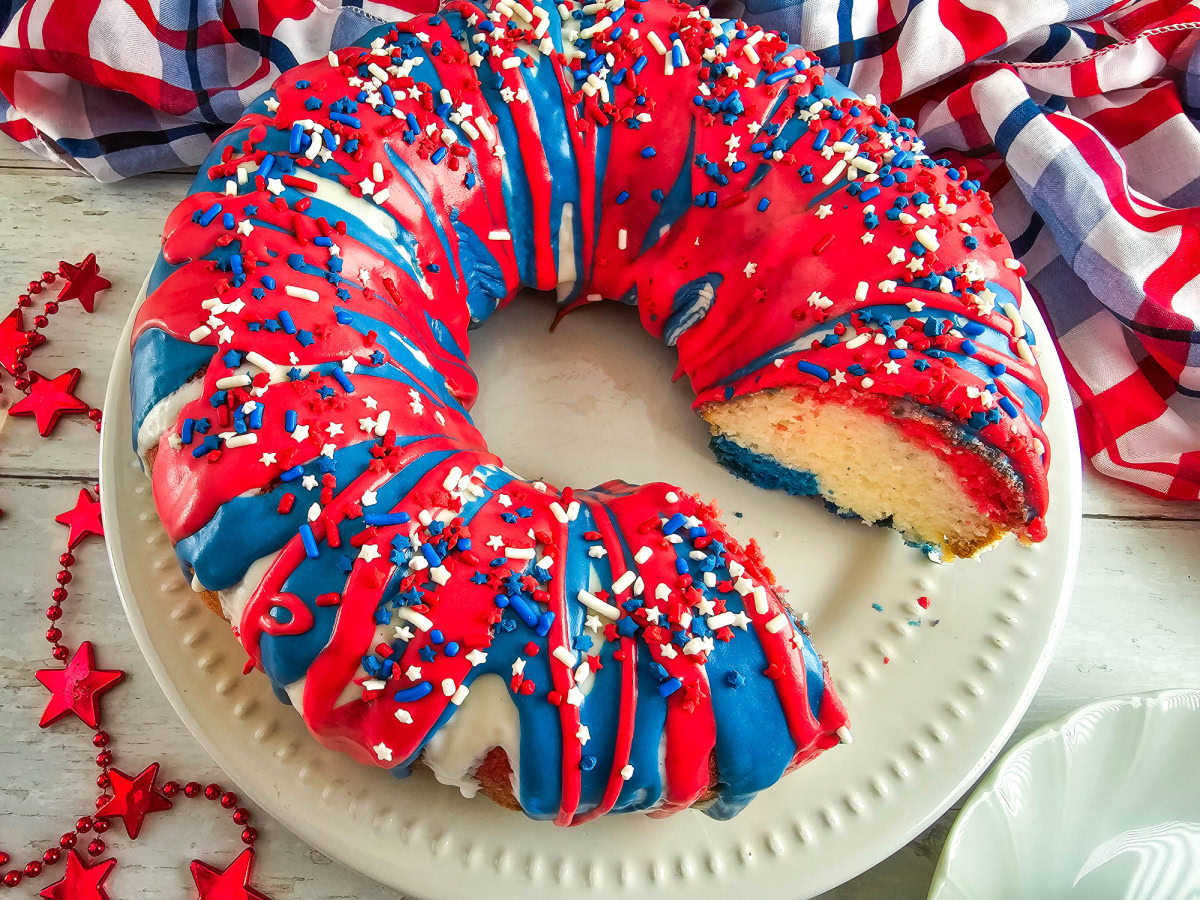 A bundt cake with red, white, and blue icing and sprinkles on a white plate, with a slice missing and a festive red, white, and blue cloth in the background.