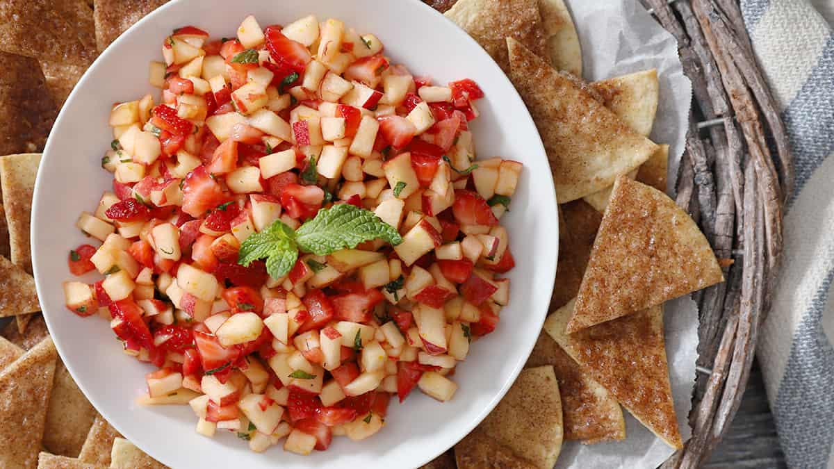 A white bowl filled with chopped apple and strawberry salsa, garnished with mint, surrounded by cinnamon-sugar tortilla chips on a platter.
