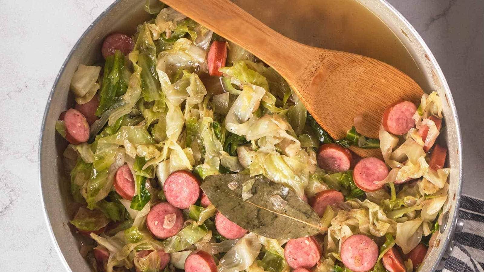 A pot of cooked cabbage and sliced sausage with carrots, broth, and a bay leaf, stirred with a wooden spoon.