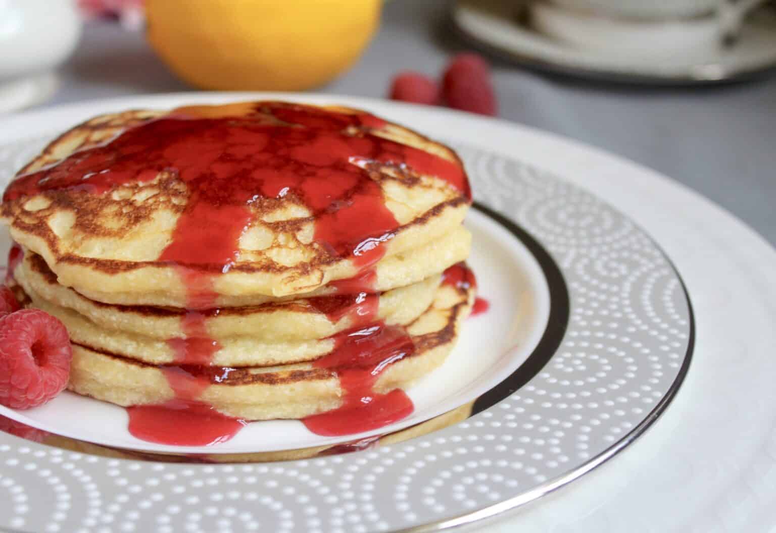 Three pancakes stacked on a decorative plate, topped with red berry syrup, with raspberries on the side.