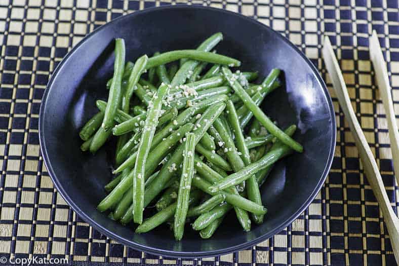 A black bowl filled with cooked green beans topped with minced garlic, placed on a black and beige woven mat with wooden chopsticks beside it.