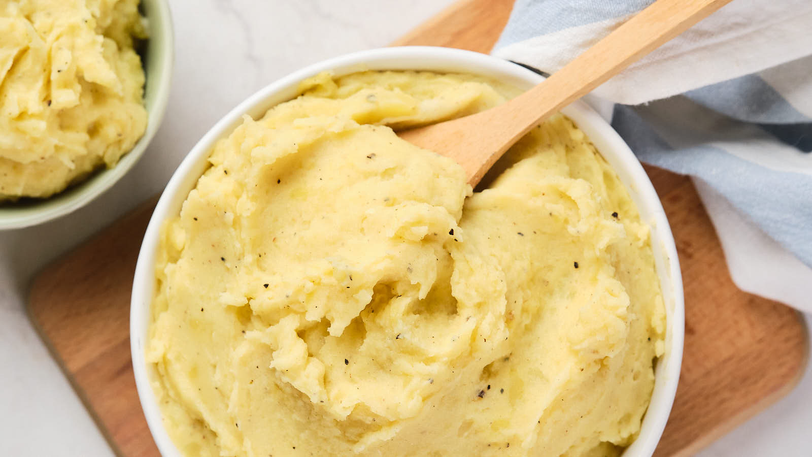 A bowl of creamy mashed potatoes with black pepper, served with a wooden spoon on a cutting board.