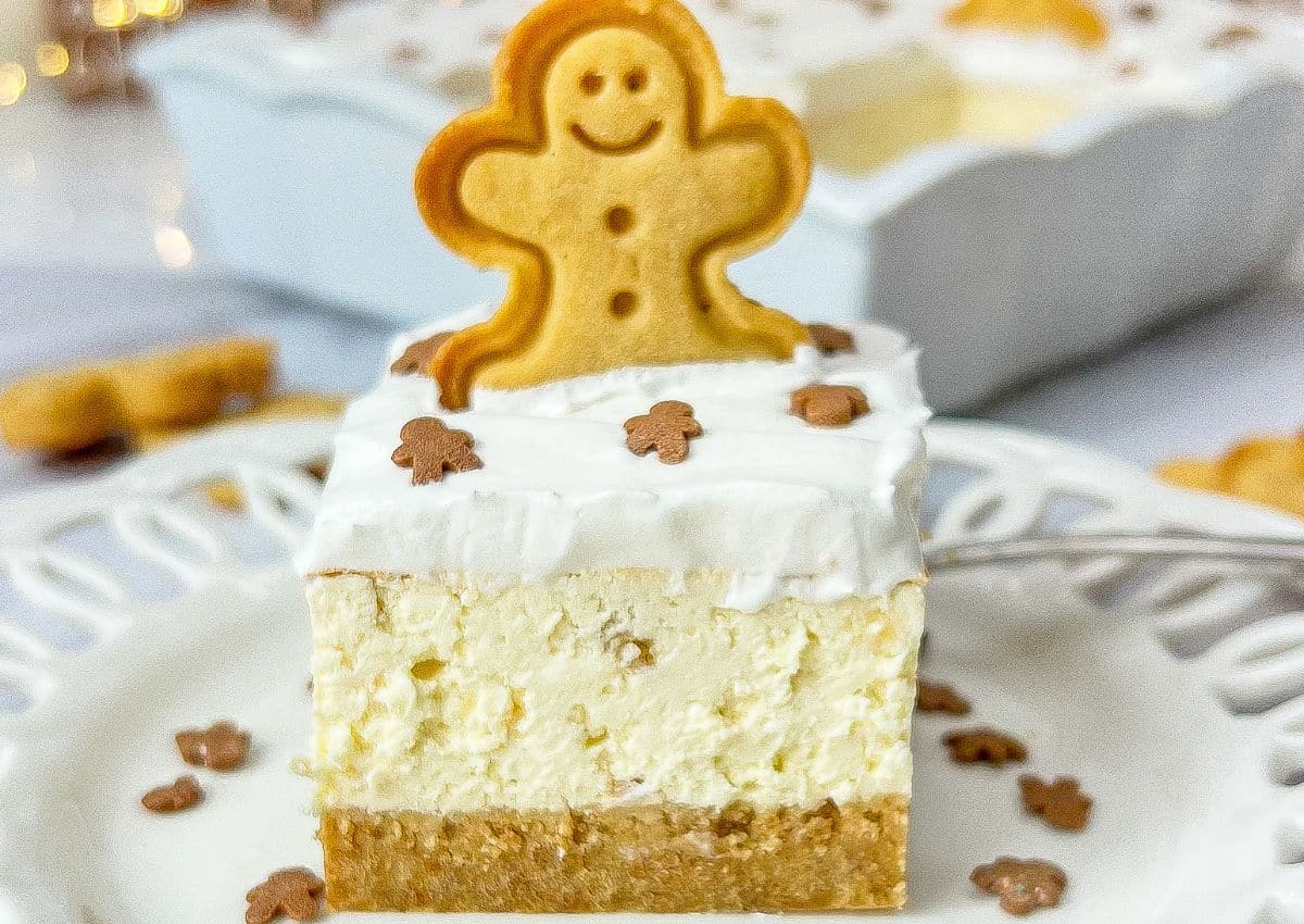 A slice of cheesecake with a graham cracker crust, topped with whipped cream, gingerbread-shaped sprinkles, and a gingerbread cookie on a white plate.