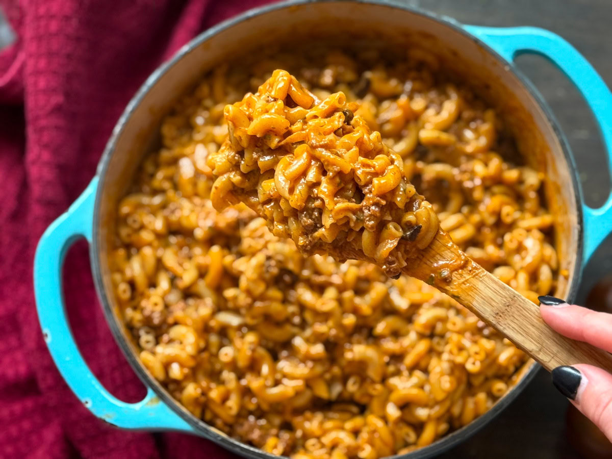 A close-up of a wooden spoon holding a serving of macaroni with ground meat in a blue pot, with a red cloth in the background.