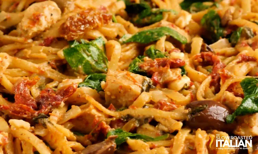 Close-up of pasta mixed with spinach, sun-dried tomatoes, olives, chicken, and creamy sauce, with The Slow Roasted Italian logo in the corner.