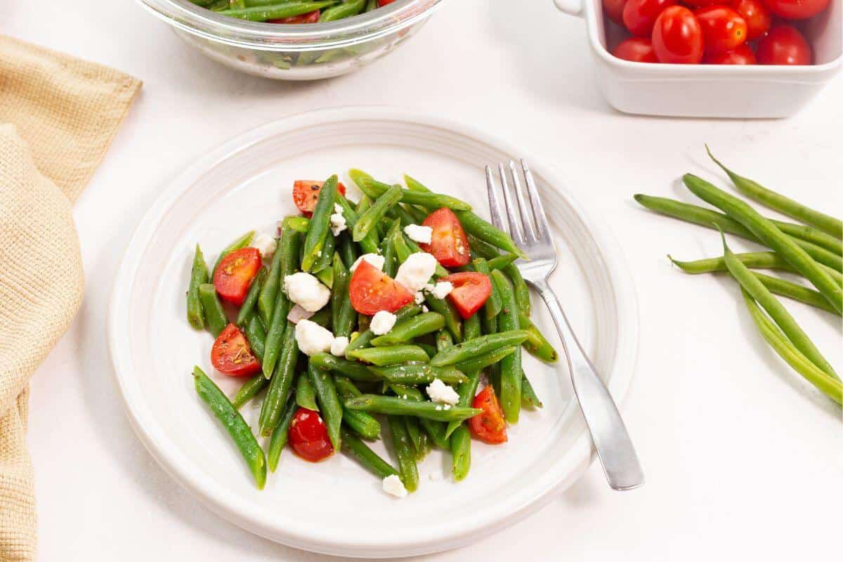 A plate of green bean salad topped with cherry tomatoes and crumbled cheese, with a fork on the side. Fresh green beans and tomatoes are in bowls nearby.