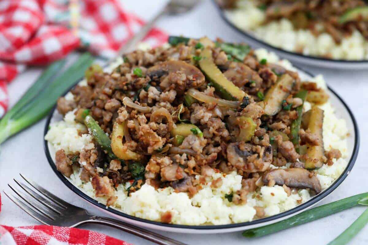 A plate of cooked ground meat with mushrooms, green vegetables, and herbs served over a bed of rice, with a fork and a red checked napkin beside it.