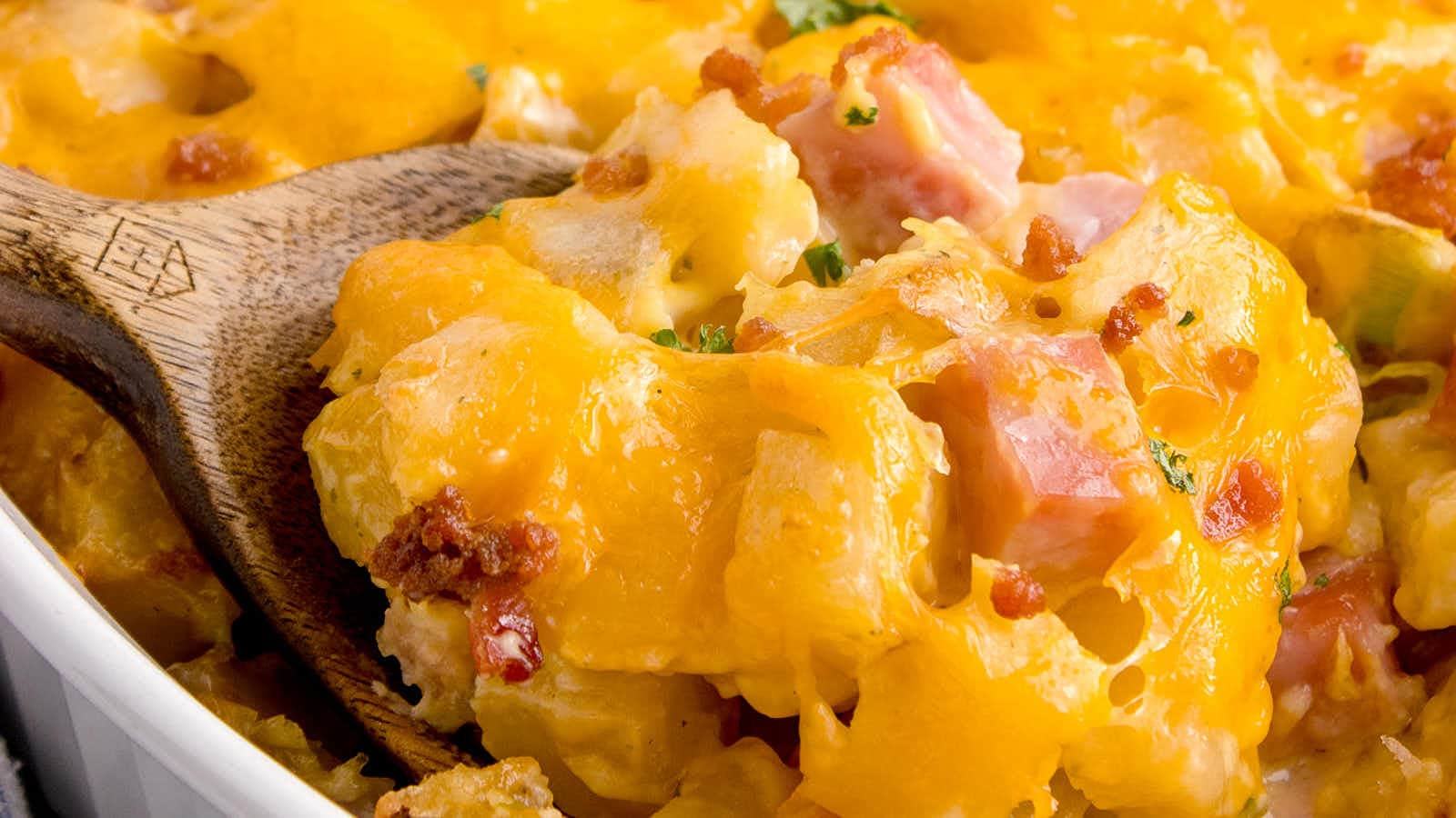 Close-up of a cheesy casserole with chunks of ham and bacon, scooped with a wooden spoon.