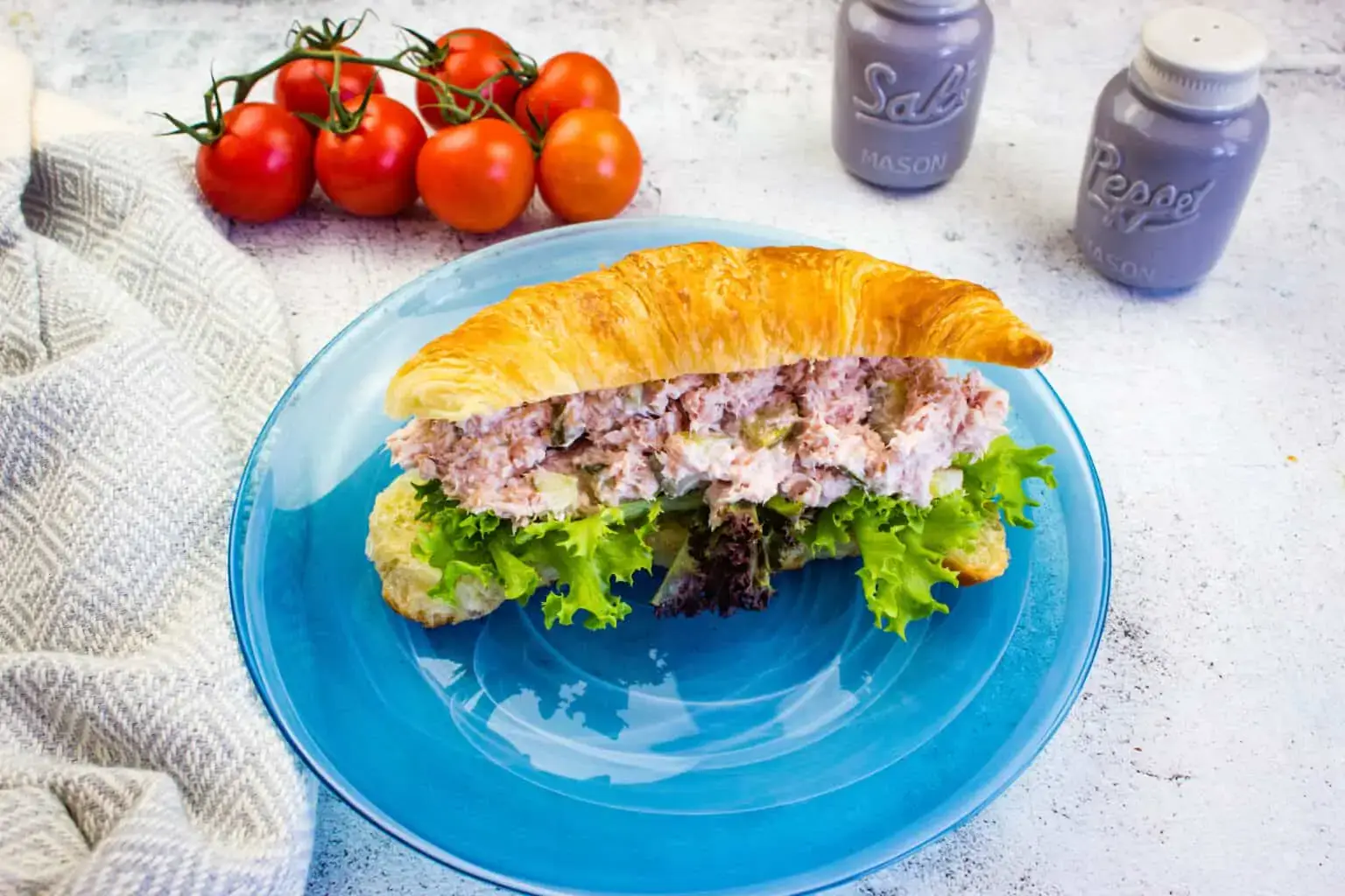 A croissant sandwich filled with tuna salad and lettuce on a blue plate, with fresh tomatoes and salt and pepper shakers in the background.