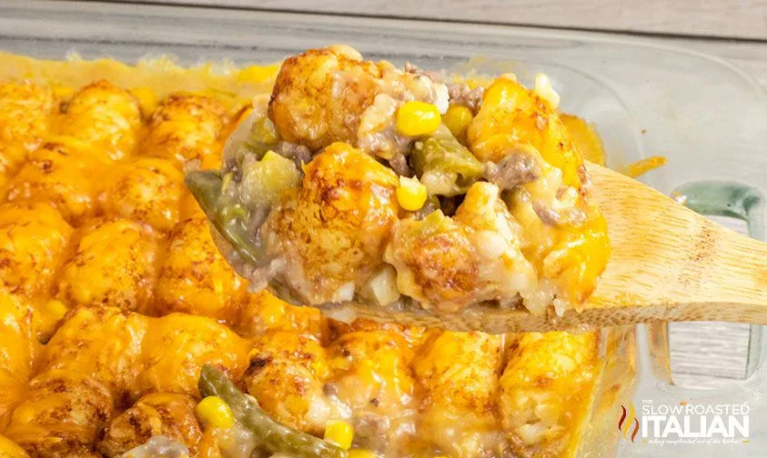 A wooden spoon lifts a serving of tater tot casserole with ground beef, cheese, corn, and green beans from a baking dish.