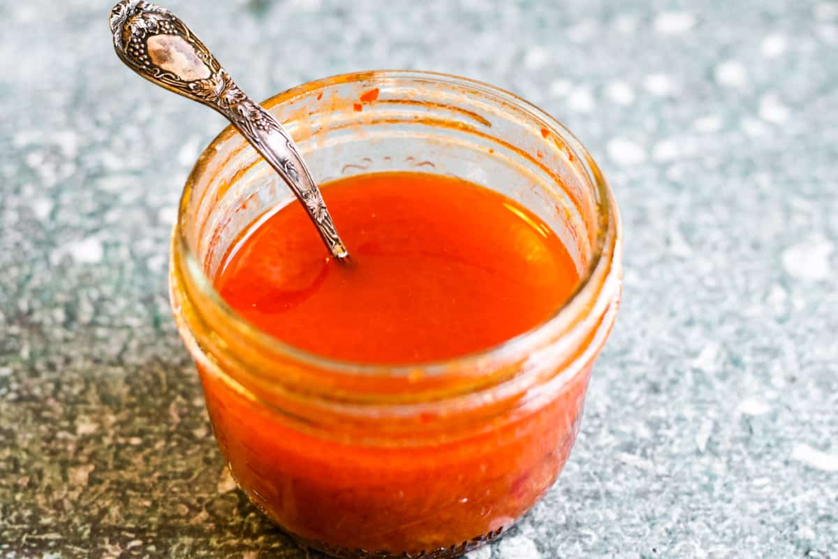 A small glass jar filled with bright orange-red sauce, with a silver spoon inside, sits on a speckled countertop.