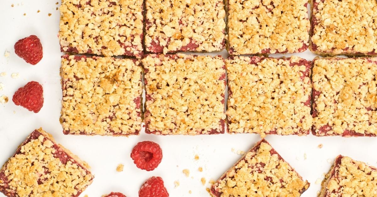 Twelve raspberry crumble bars arranged in rows on a white surface, with a few fresh raspberries scattered around.