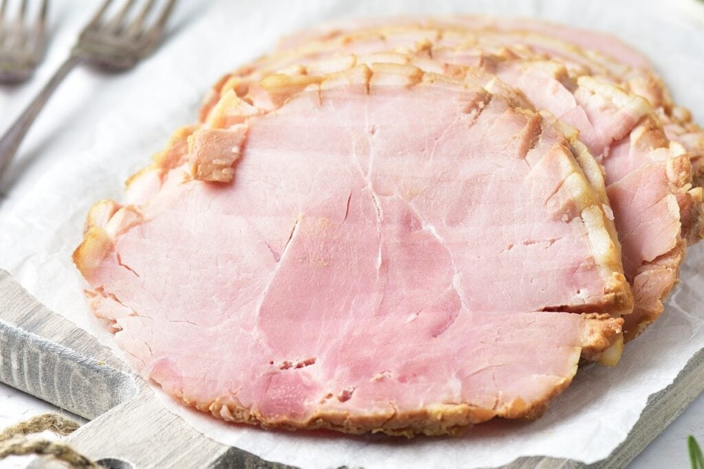 Several slices of cooked ham are arranged on a white parchment paper atop a wooden cutting board, with two metal forks in the background.