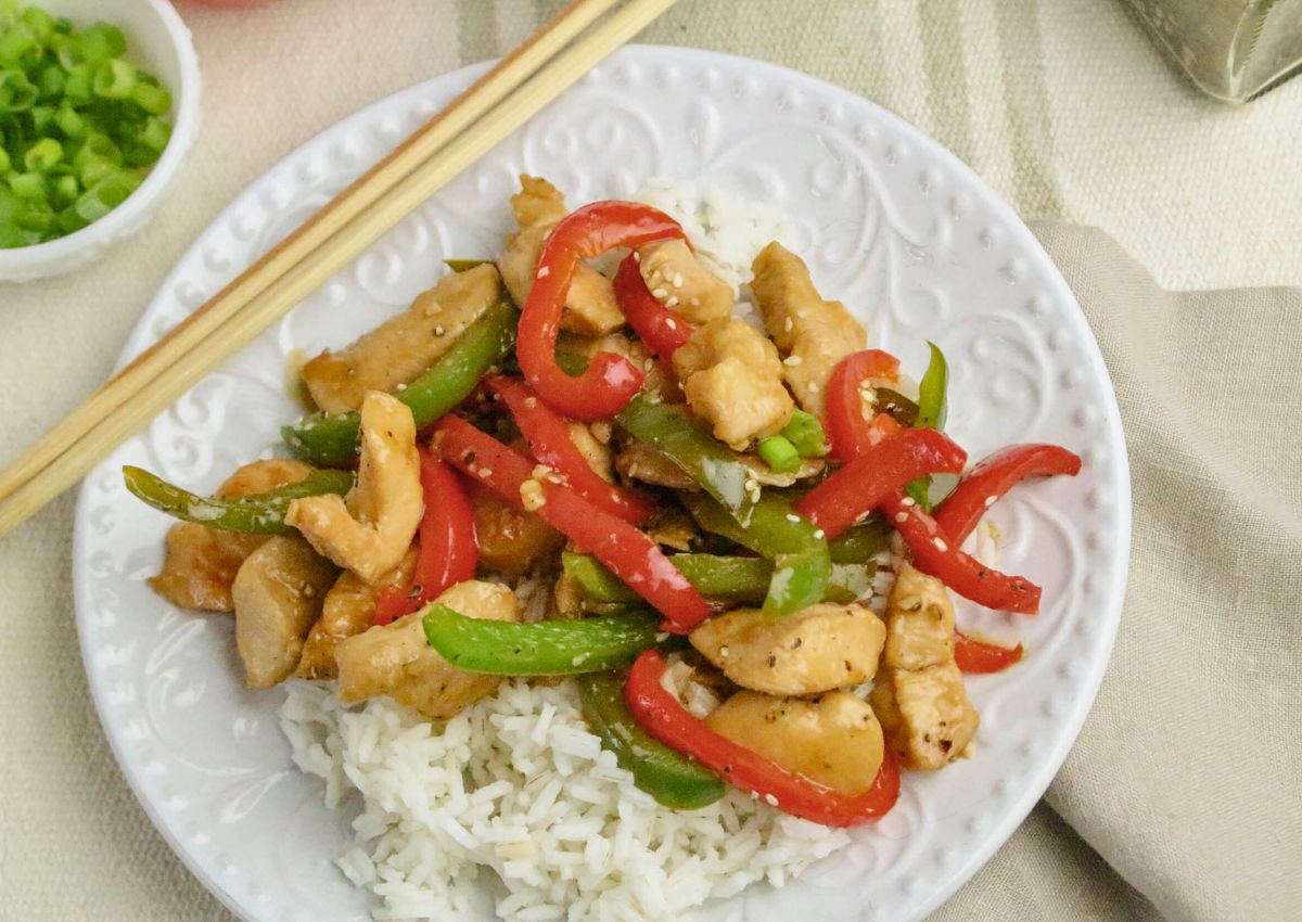 A plate of white rice topped with stir-fried chicken strips, red and green bell peppers, and sauce, with chopsticks resting on the plate.