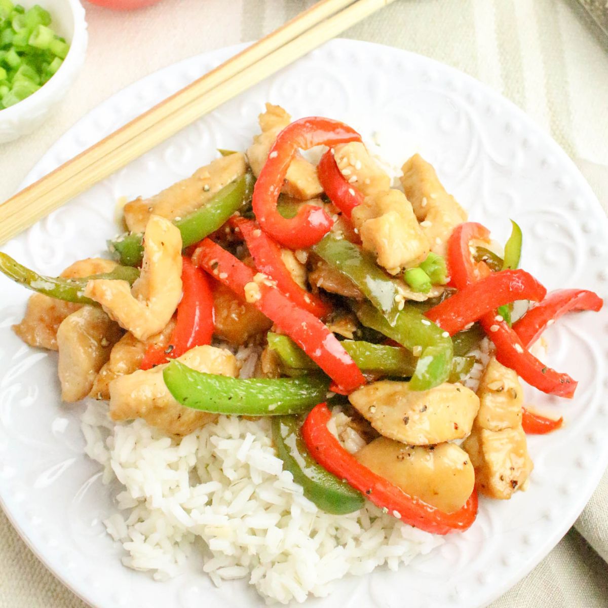 A plate of white rice topped with stir-fried chicken, red and green bell peppers, and sesame seeds, served with chopsticks.
