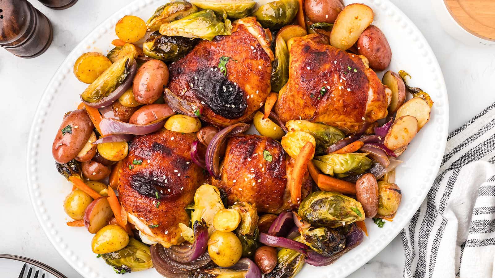A white plate with three baked chicken thighs surrounded by roasted potatoes, Brussels sprouts, carrots, and red onions.