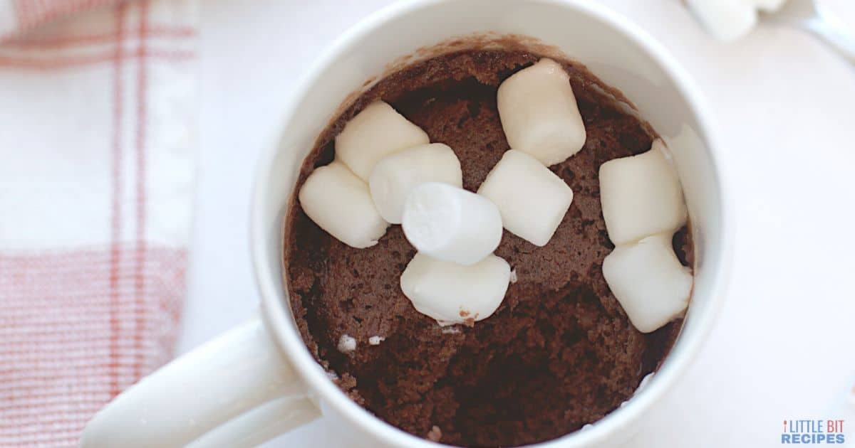 A white mug filled with chocolate mug cake topped with mini marshmallows, placed on a white surface next to a red and white towel.