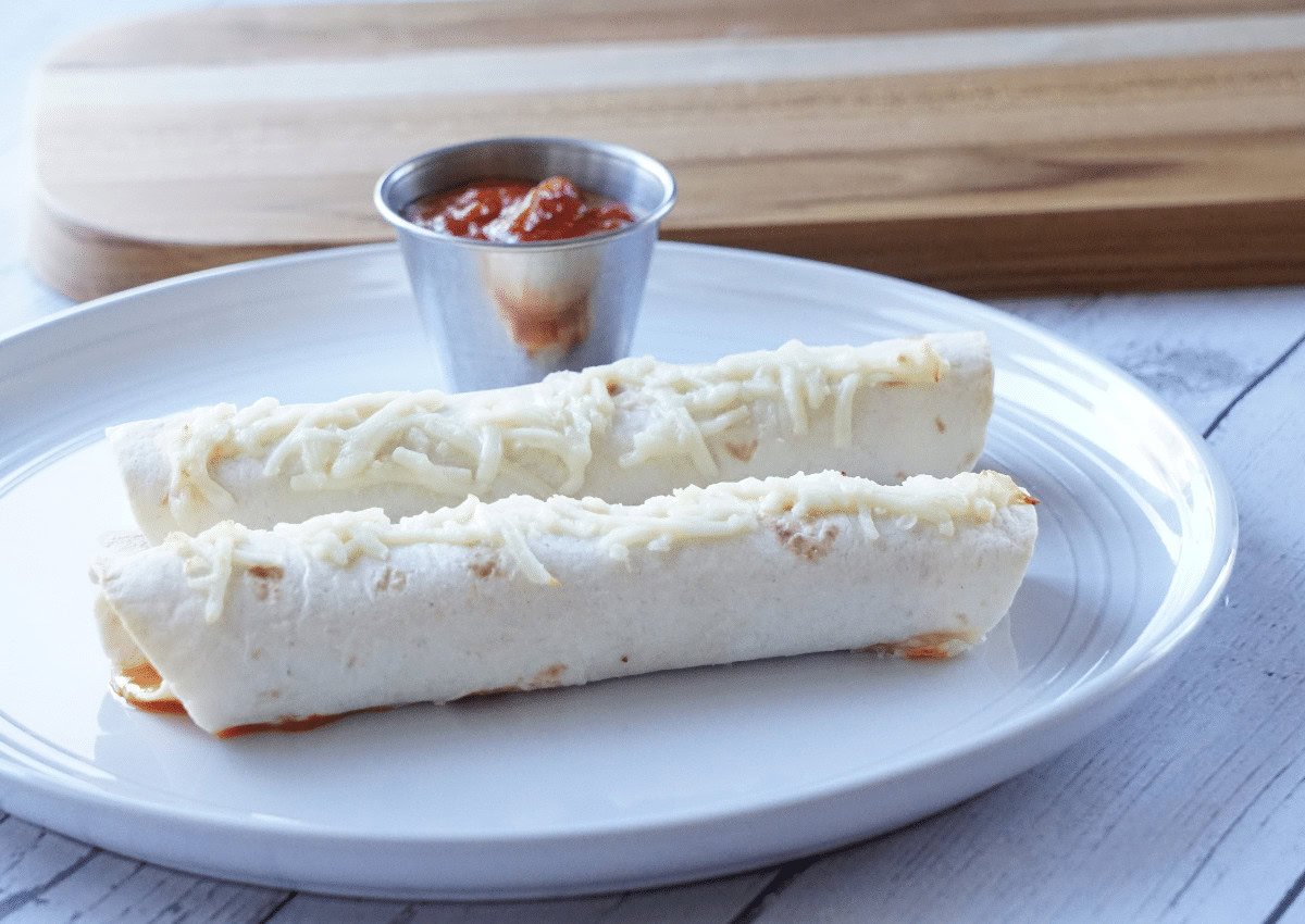 Two cheese-topped rolled tortillas are served on a white plate with a small metal cup of salsa, set on a light wooden table.