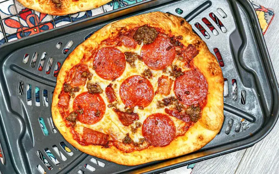 A baked pizza with pepperoni, sausage, and bacon toppings sits on a black perforated pizza pan, placed on a patterned cloth.