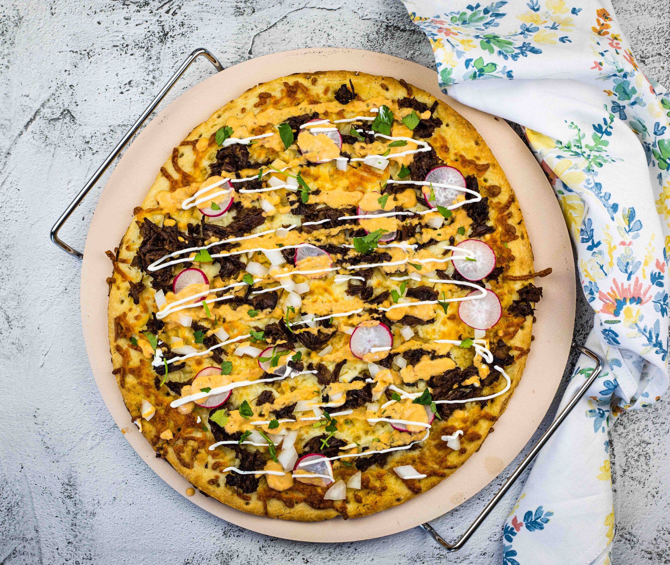 A pizza topped with cheese, ground beef, sliced radishes, onions, cilantro, and drizzled with a white sauce, served on a round baking tray next to a floral-patterned napkin.