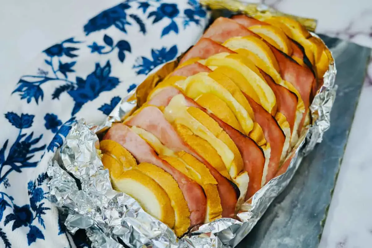 Loaf pan lined with foil, filled with alternating slices of bread, ham, and cheese, placed next to a white and blue-patterned cloth.