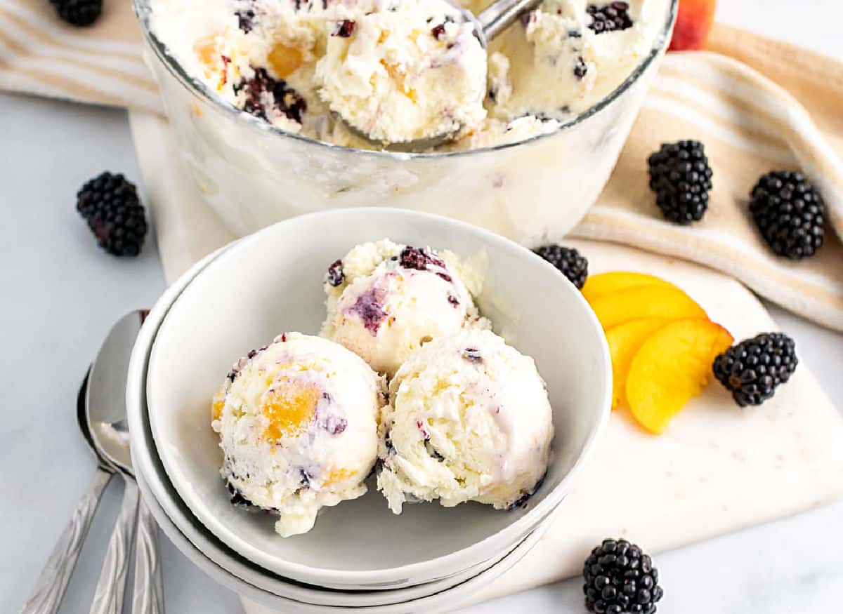 Three scoops of ice cream with blackberries and peach chunks in a bowl, with fresh blackberries and peach slices nearby and a glass bowl of ice cream in the background.