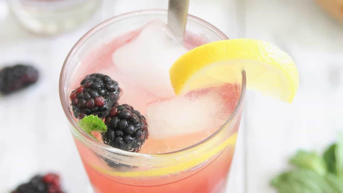 A glass of pink lemonade with ice cubes, blackberries, a lemon slice, and a mint garnish, with a metal straw.