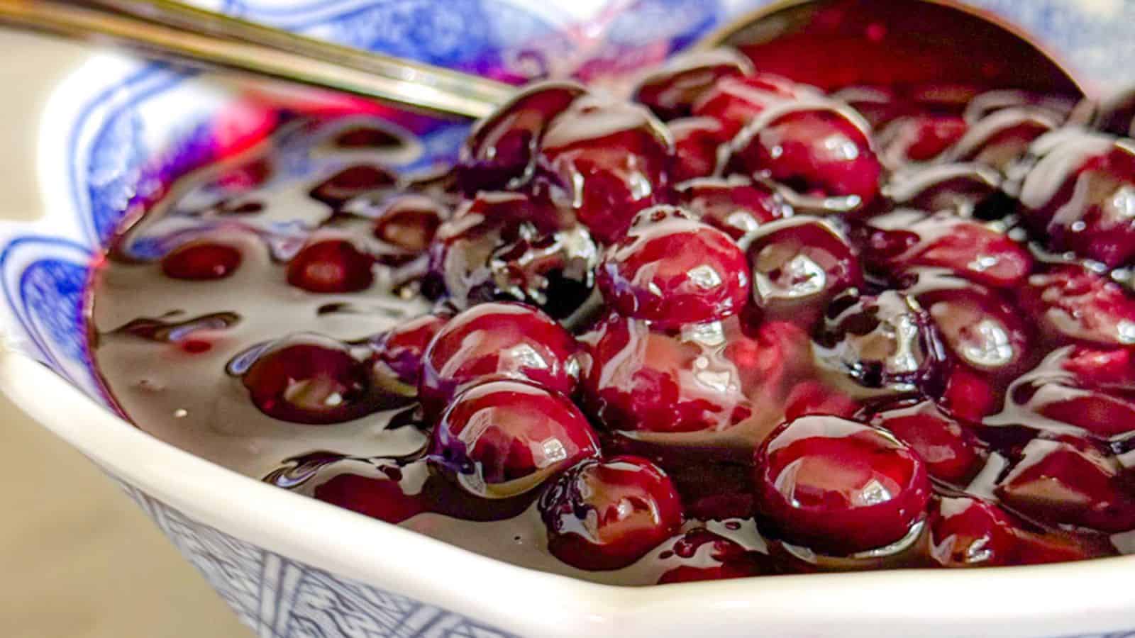 Close-up of a bowl filled with glossy, cooked blueberries in a thick syrup, with a spoon partially visible in the background.