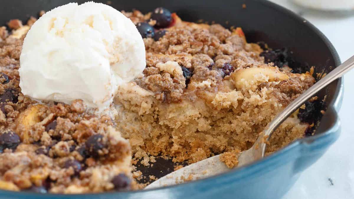 A fruit crumble with a crumb topping and a scoop of vanilla ice cream in a blue skillet, with a portion served out and a spoon in the dish.