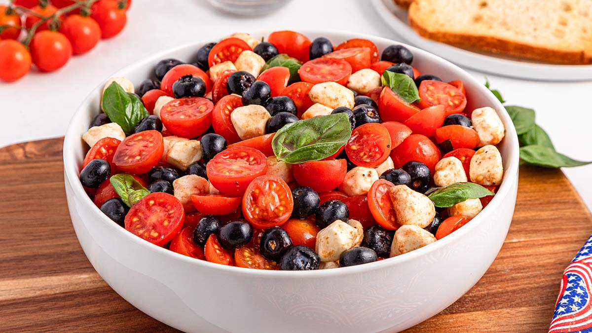 A white bowl filled with cherry tomatoes, black olives, mozzarella balls, and fresh basil leaves sits on a wooden surface.