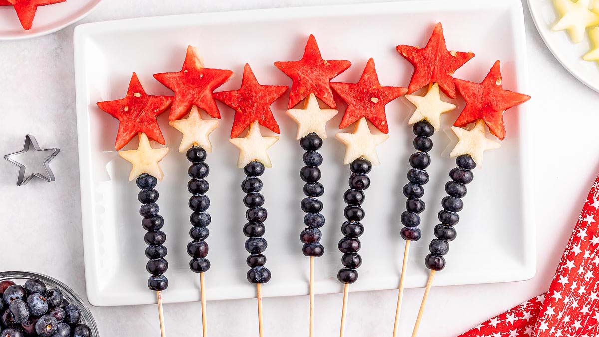 Skewers with watermelon stars, white melon stars, and blueberries arranged on a white plate, resembling patriotic wands.