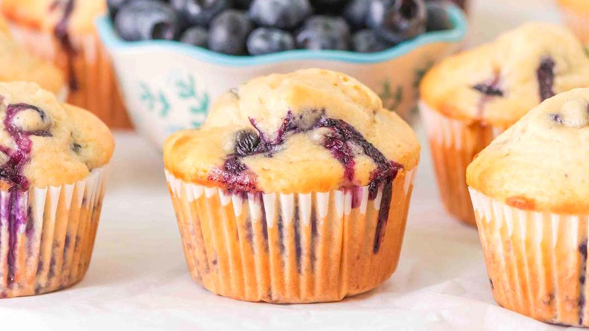 Several blueberry muffins are arranged on a light surface, with a bowl of fresh blueberries in the background.