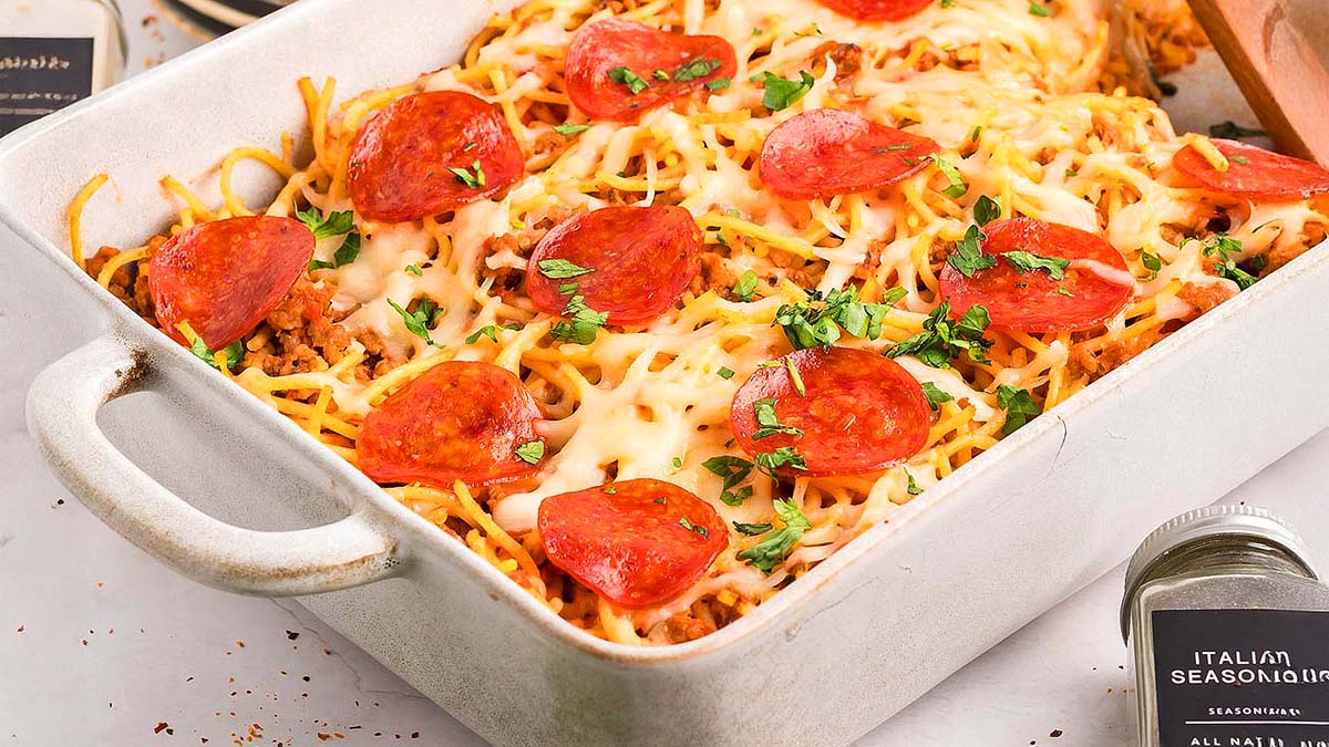 A rectangular casserole dish filled with baked spaghetti topped with melted cheese, sliced pepperoni, and chopped herbs sits on a table.