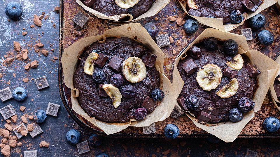 Chocolate muffins with banana slices and blueberries in parchment paper, in a muffin tray. Blueberries and chocolate chunks scattered on a dark surface around the muffins.