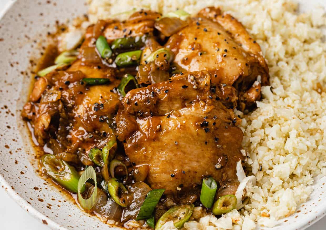 A plate of glazed chicken thighs topped with sliced green onions and black pepper, served with a side of cauliflower rice.