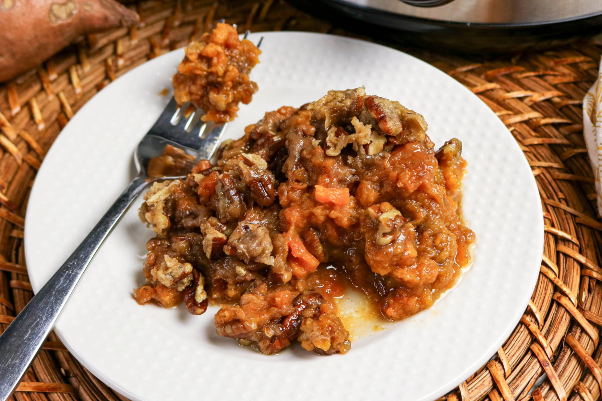 A serving of sweet potato casserole with pecans is on a white plate with a fork, placed on a woven surface.