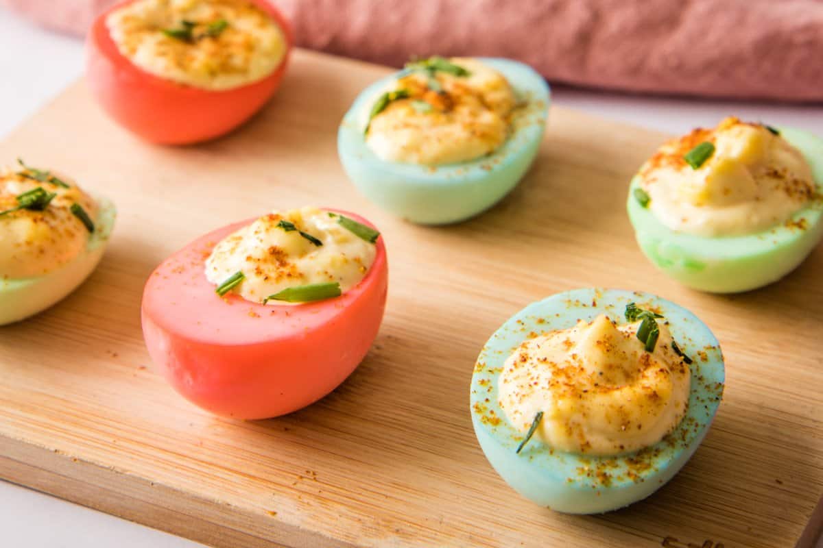 Three pink and three blue deviled eggs topped with chives and paprika are arranged on a wooden board.