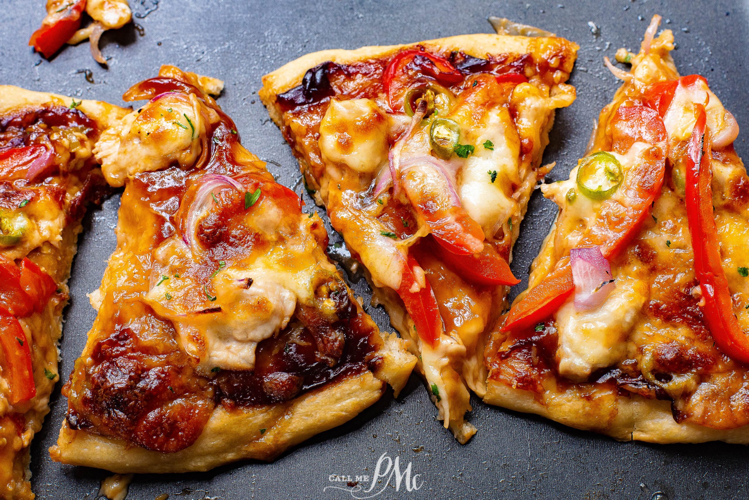 Four slices of pizza topped with cheese, red bell peppers, onions, jalapeรฑos, and barbecue sauce on a dark surface.