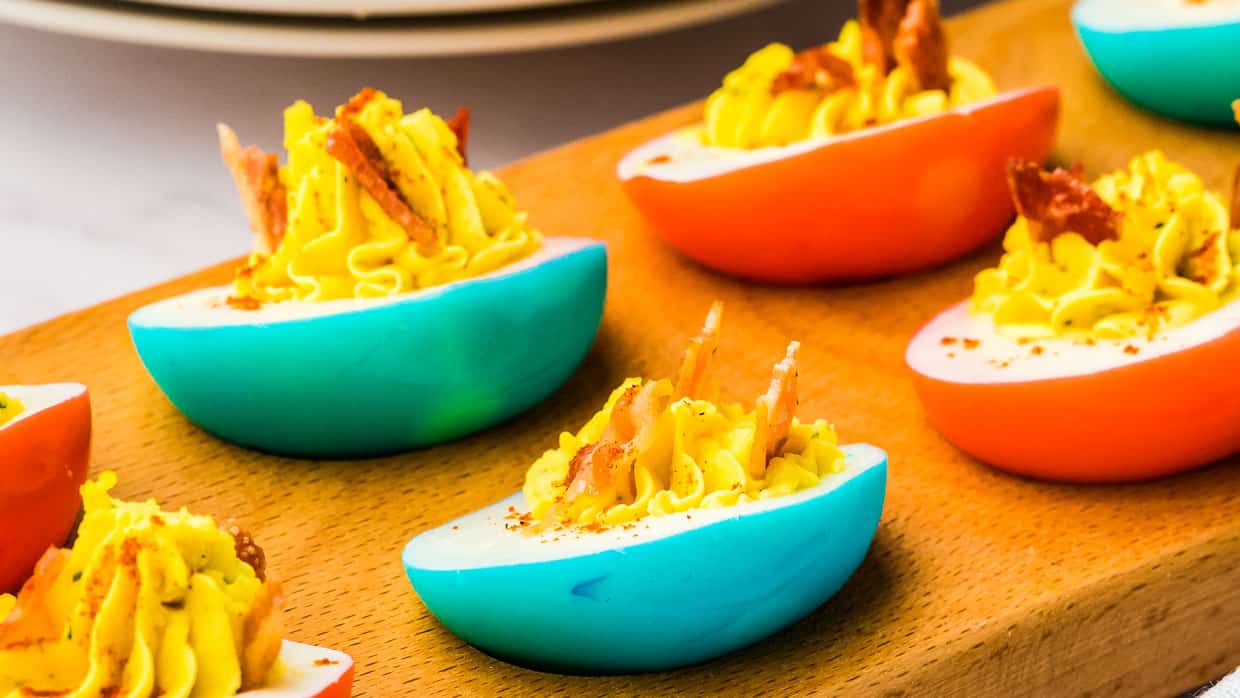Deviled eggs with yellow filling are served in brightly colored blue and orange egg white halves, garnished with small bacon pieces, on a wooden tray.