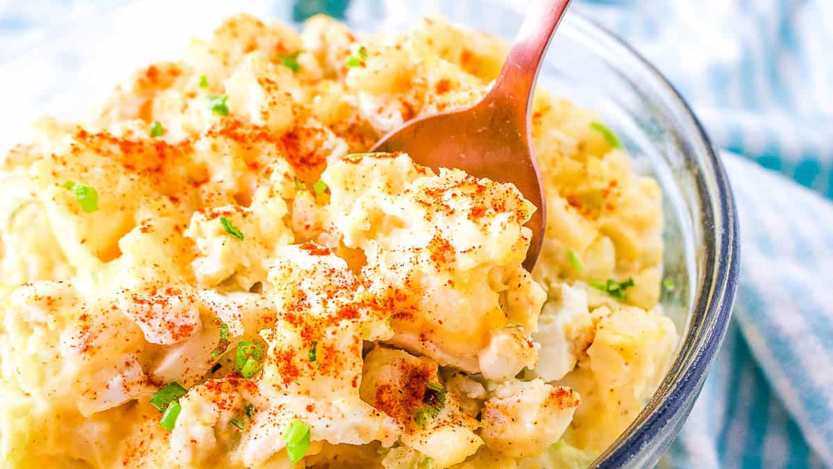 A bowl of creamy potato salad topped with paprika and chopped green onions sits on a table with a spoon inside.